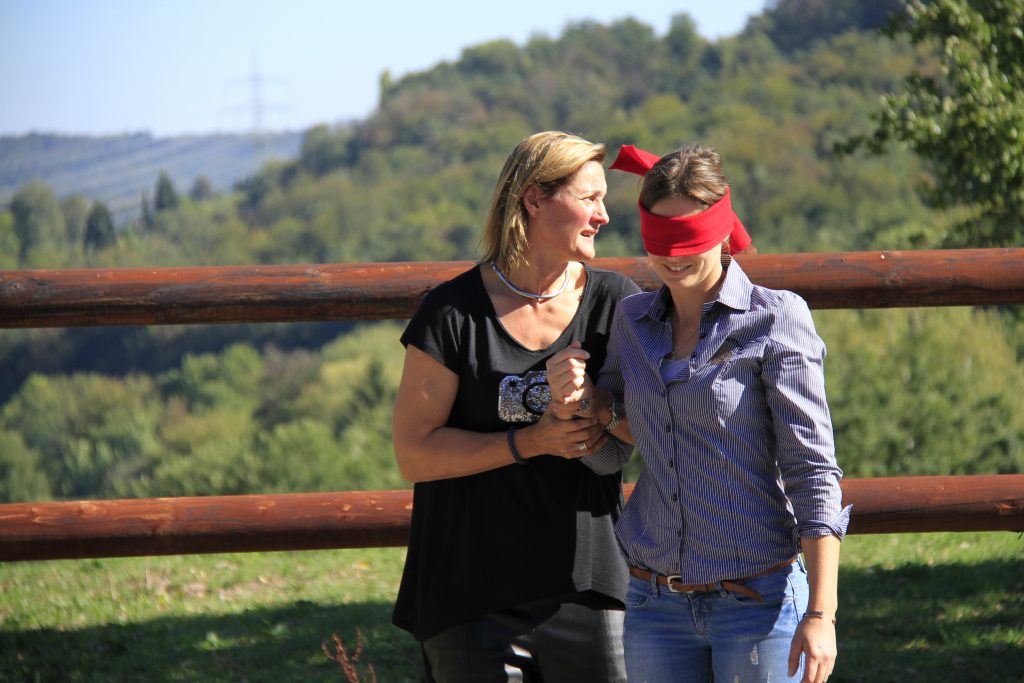 Weibliche Person mit roter Augenbinde wird von weiterer weiblicher Person geführt. Im Hintergrund Pferdekoppel und Bäume.